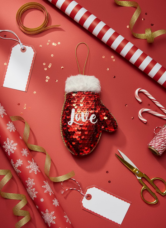 Red sequin mitten ornament with 'Love' on a red background with gift wrap, ribbons, and scissors.
